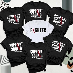May include: Six black t-shirts with white text and a red ribbon design. The text on the shirts reads "Support Squad #TeamEmma" and "Fighter".