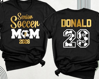 Peut inclure: Un t-shirt noir avec du texte doré et blanc indiquant Senior Soccer MOM 2026 et un motif de ballon de football. Le dos du t-shirt affiche le nom DONALD en doré au-dessus du numéro 29, rempli d'un motif de ballons de football.