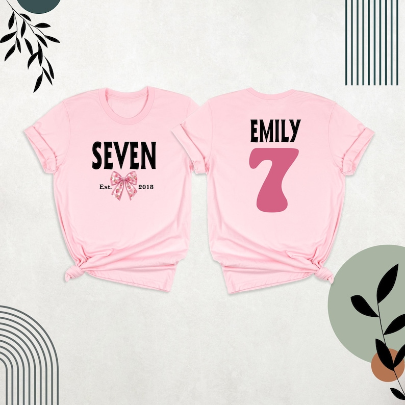 Peut inclure: Deux t-shirts roses, l'un avec le texte "SEVEN Est. 2018" et un n&oelig;ud rose, l'autre avec le texte "EMILY" et un grand chiffre 7 rose.