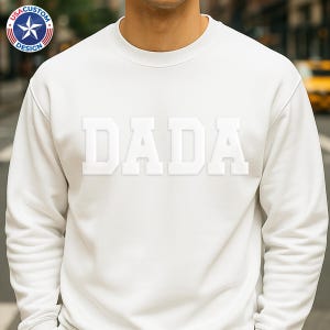 May include: A white sweatshirt with the word "DADA" in bold, raised, white letters across the chest. The sweatshirt has a crew neck and long sleeves. A logo with a star and the words "USA CUSTOM DESIGN" is visible in the upper left corner.