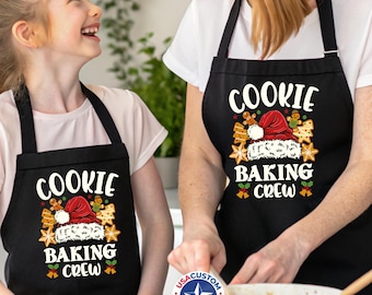May include: Black aprons with the text "COOKIE BAKING CREW" in white, red, and green. The design includes a Santa hat, gingerbread men, and star-shaped cookies. The aprons are worn by two people.