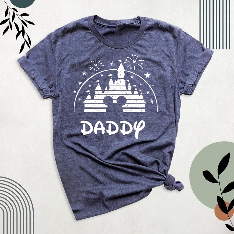 May include: A navy blue t-shirt with a white graphic of a castle, fireworks, and stars. The word "DADDY" is printed in a stylized font below the castle design. The shirt is knotted at the bottom.