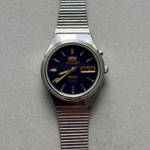 May include: A silver-toned Orient watch with a dark blue face and a silver band. The watch face displays gold hands and markers, with the day and date visible. The watch has a classic, retro design.