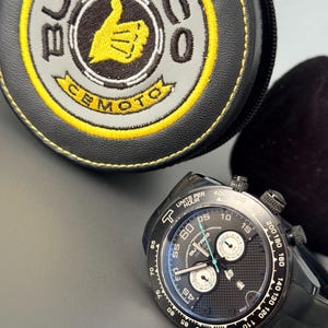 May include: Black wristwatch with a black dial and a black rubber strap. The watch has white and blue accents and multiple sub-dials. A black and yellow Bultaco case is in the background.