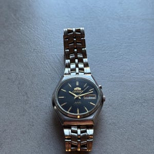 May include: A silver Orient watch with a black face and a silver metal band. The watch has a date window and the words "Orient" and "ARA" printed on the face.