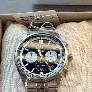Op de afbeelding: Een zilverkleurig Seiko chronograaf horloge met een zwarte wijzerplaat en drie witte sub-wijzerplaten. Het horloge heeft een roestvrijstalen schakelarmband, een tachymeter-ring en lichtgevende wijzers. Een label toont Ref. SSB427P1.