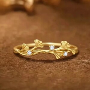 May include: A gold-colored ring with a delicate, nature-inspired design. The ring features a flowing vine-like structure with three small, clear gemstones and stylized leaf accents. The ring is set against a warm brown background.