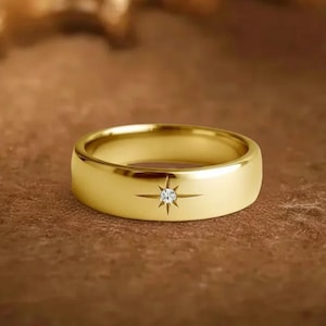 Può includere: Un anello d'oro con un design a stella e una piccola gemma trasparente al centro. L'anello è posto su uno sfondo marrone, con un elemento decorativo dorato sfocato sullo sfondo.