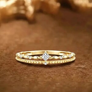 Op de afbeelding: Een goudkleurige ring met een dubbel bandontwerp. De bovenste band heeft een vierkant geslepen diamant als middensteen, met kleinere stenen en decoratieve elementen. De ring is geplaatst tegen een bruine achtergrond, die de ingewikkelde details benadrukt.