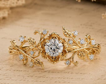 May include: A gold ring featuring a central round brilliant-cut clear gemstone set within a floral-shaped mounting, surrounded by intricate gold vine and leaf motifs accented with small round clear gemstones along the band's branches.