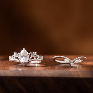May include: Two silver rings with clear gemstones. The larger ring has a pear-shaped stone and a crown-like design. The smaller ring has a V-shaped design with three smaller stones.