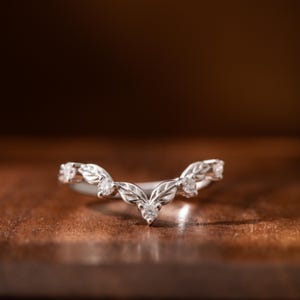 Peut inclure: Une bague en argent avec un motif de feuilles et de petites pierres précieuses transparentes. La bague a une forme incurvée, avec les feuilles et les pierres précieuses disposées le long de la courbe. Fond brun chaud.