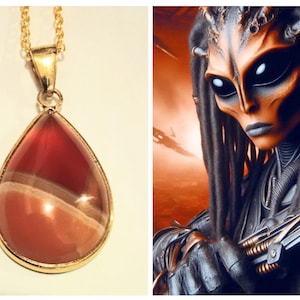 May include: A gold-colored necklace with a teardrop-shaped pendant. The pendant features a polished, layered stone in shades of red, brown, and cream. Next to it is a digital art image of an alien with dark dreadlocks and a metallic suit.