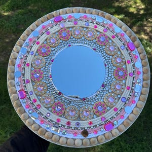Puede incluir: Un espejo redondo de madera con un diseño rosa y blanco. El espejo está decorado con diamantes de imitación, cuentas y pequeños círculos de madera.