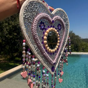 Könnte beinhalten: Herzförmiger Wandschmuck mit zentralem Spiegel, silberfarbenen Verzierungen und einem lavendelfarbenen Rand. Er hat herabhängende Stränge aus Perlen, Perlen und rosa herzförmigen Charms. Das Stück wird von einer Hand gehalten.