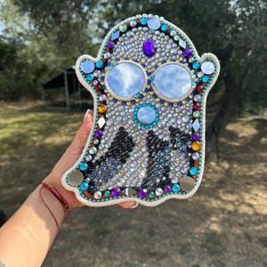 Puede incluir: Un adorno de pared con forma de fantasma blanco decorado con brillantes de colores y ojos de espejo.