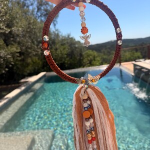 Puede incluir: Un atrapasueños de macramé marrón con cristales transparentes y naranjas, un dije de mariposa plateada y una borla de hilo blanco y naranja. Cuentas decorativas y un pequeño dije de ángel cuelgan del centro. El atrapasueños se sostiene contra un cielo azul.