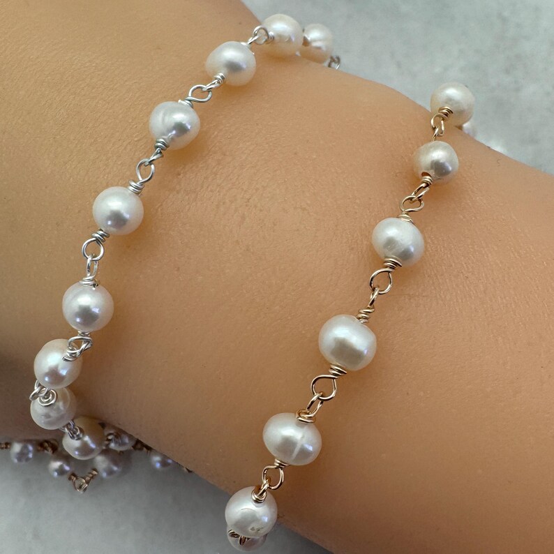 Puede incluir: Una delicada pulsera con brillantes perlas blancas ensartadas en alambre plateado y dorado. Las perlas est&aacute;n espaciadas uniformemente, creando un dise&ntilde;o cl&aacute;sico y elegante. La pulsera se muestra en una mu&ntilde;eca.