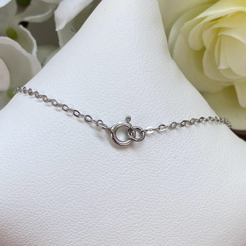 May include: A delicate silver chain necklace with a spring ring clasp. The necklace is displayed on a white surface, with a blurred floral background. The chain is made of small, interconnected links.
