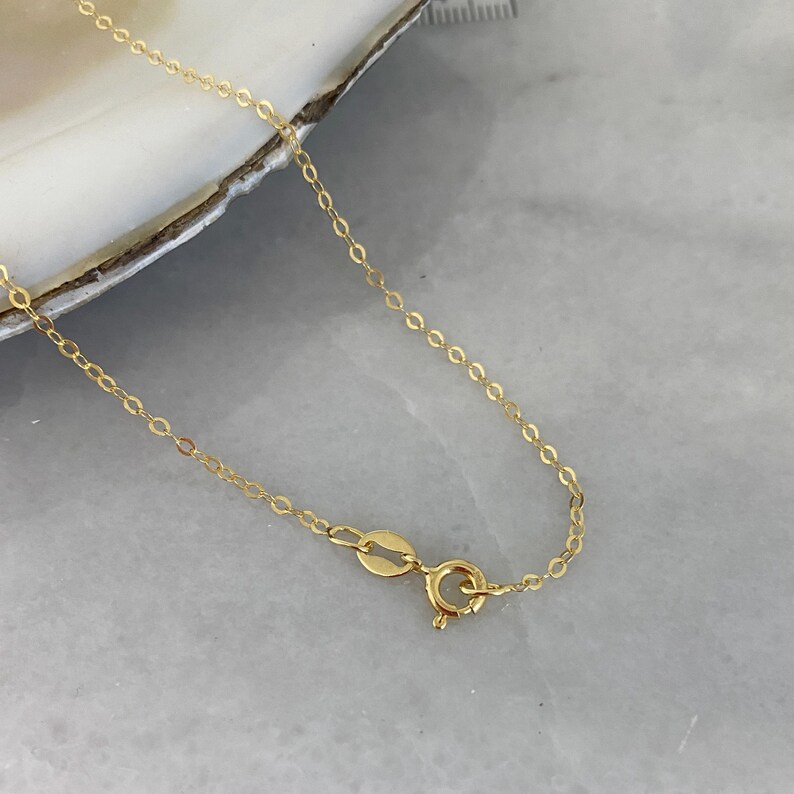 May include: A delicate gold chain necklace featuring thin, interlocking oval links and a secure spring ring clasp. The metal has a polished, reflective finish, and the chain is shown in its entirety with the clasp mechanism clearly visible.