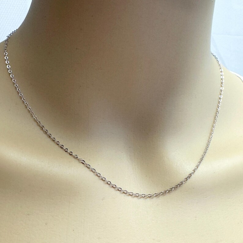 May include: A delicate silver chain necklace with small, evenly spaced links. The necklace is displayed on a mannequin, showcasing its simple and elegant design. The chain is a classic accessory.