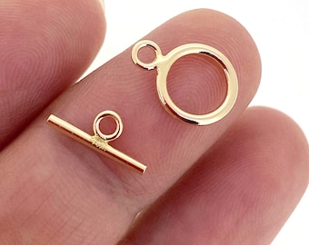 May include: Close-up of two gold-colored jewelry clasps. One is a small, straight bar with a loop, and the other is a larger circular ring with a loop. The clasps are likely for necklaces or bracelets.