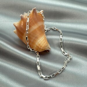 May include: A silver chain necklace with a box chain design. The chain is lying on a gray fabric background with a seashell.