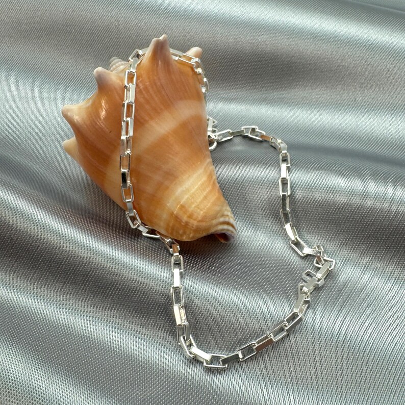 May include: A silver chain necklace with a box chain design. The chain is lying on a gray fabric background with a seashell.