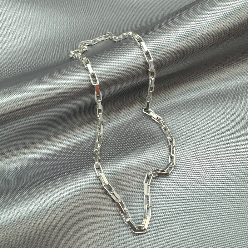 May include: A silver chain necklace with a box chain design. The chain is lying on a gray fabric background.