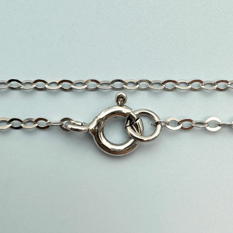 May include: Close-up of a silver-colored necklace with a lobster clasp. The chain is made of small, oval links. The clasp is round with a lever for secure closure. The background is a light blue.