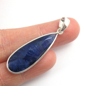 May include: A teardrop-shaped pendant with a faceted, deep blue gemstone set in a silver-colored metal frame. The pendant is held against a light background, showcasing its polished surface and intricate design. The gemstone's color varies from dark to light blue.