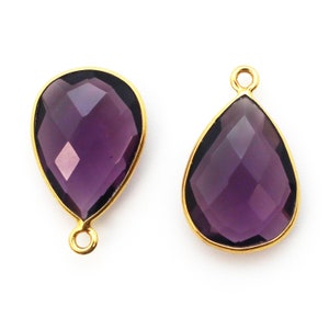 May include: Two gold-plated teardrop-shaped pendants with purple faceted glass stones. The pendants have a loop at the top for attaching to a chain or cord.