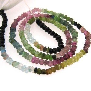 May include: Strands of colorful tourmaline beads in shades of black, pink, green, blue, and yellow. The beads are small, faceted, and strung together, creating a vibrant and eye-catching display. The beads are on a white background.