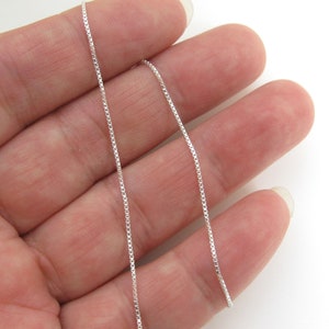 May include: A silver chain necklace with a box chain design. The chain is thin and delicate, and it is shown on a hand.