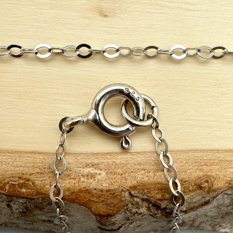 May include: Close-up of a silver necklace with a circular clasp and delicate chain links. The clasp is marked with "925". The necklace is displayed on a wooden surface, showcasing its fine details and craftsmanship.