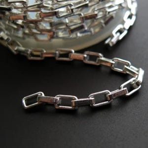 Puede incluir: Cadena de plata con eslabones cuadrados. La cadena está enrollada en un carrete y una sección de la cadena está extendida.