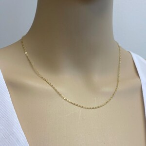May include: A delicate gold chain necklace featuring small, uniform oval links. The chain has a polished, reflective finish and rests against the neck, showcasing a simple, continuous design without any additional pendants or decorative elements.