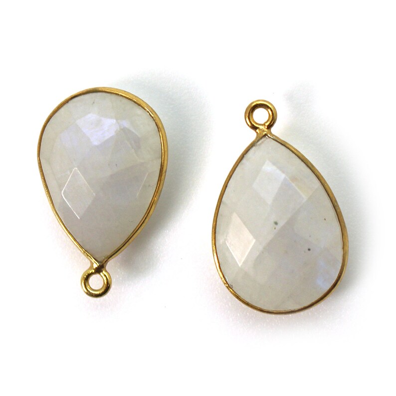 May include: Two gold-toned teardrop-shaped pendants with a white moonstone gemstone. Each pendant has a loop at the top for attaching to a chain or cord.