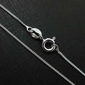 May include: A close-up of a silver curb chain necklace with a lobster clasp. The chain is made of small, interlocking links and has a shiny, reflective surface. The clasp is also silver and has a secure closure. The background is black.