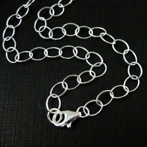 May include: A silver-colored necklace with oval-shaped links. The chain is connected with a lobster clasp. The necklace is displayed on a black surface.