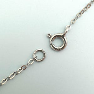 May include: Close-up of a silver necklace chain with a spring ring clasp and a small circular ring. The chain is made of small, oval-shaped links. The clasp is open, showing the details of the mechanism.