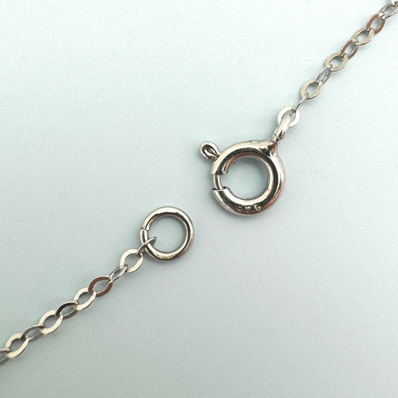 May include: Close-up of a silver necklace chain with a spring ring clasp and a small circular ring. The chain is made of small, oval-shaped links. The clasp is open, showing the details of the mechanism.