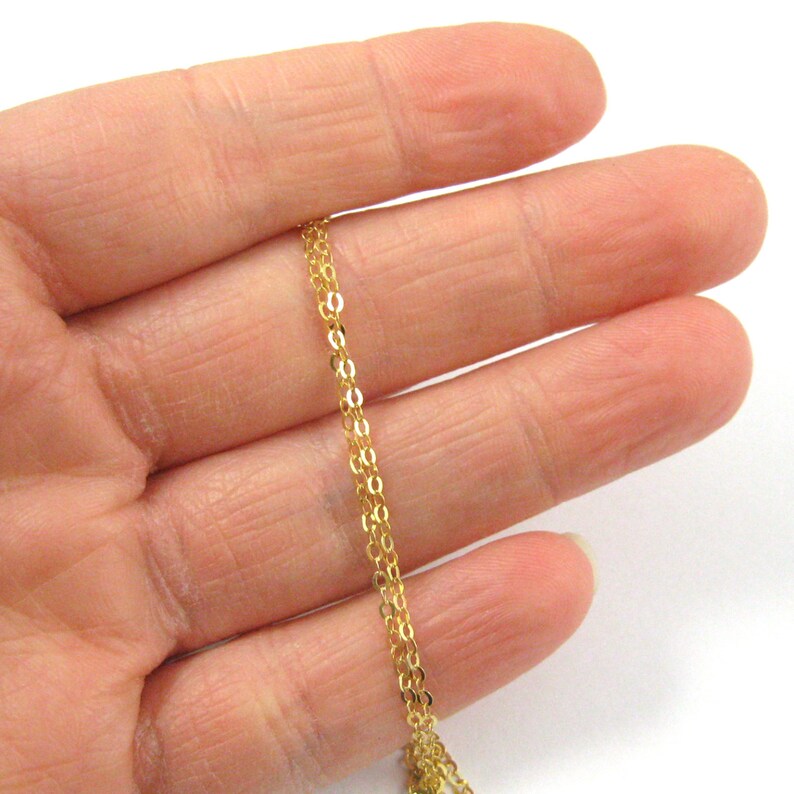 May include: A gold chain necklace with a delicate, fine link design. The chain is shown draped over a hand, showcasing its length and the intricate detail of the links.