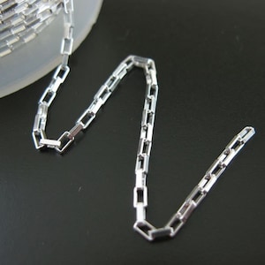 May include: A close-up of a silver-colored rectangular link chain. The chain is displayed against a dark background, with a portion of the chain coiled on a white surface. The links are uniform in size and shape.