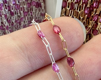 May include: Close-up of two delicate chains. One is silver-toned, the other gold-toned. Both feature oval, magenta-colored gemstones set between rectangular links. The background shows more chain, creating a sense of abundance and variety.
