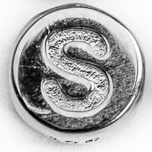 May include: A close-up of a silver-toned metal button with the letter "S" embossed on it.