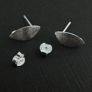 May include: A pair of silver stud earrings with a textured, leaf-like design. The earrings are shown with two earring backs. The earrings have a post for insertion into the ear. The earrings are on a dark surface.