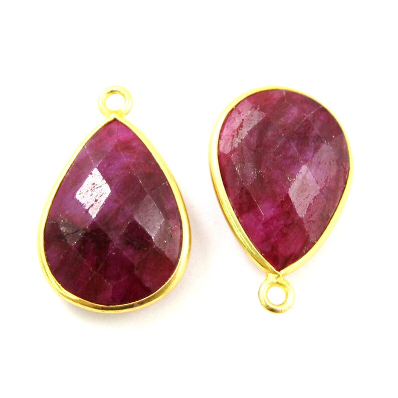 May include: Two gold-plated teardrop-shaped pendants with red ruby gemstones. The pendants have a loop at the top for attaching to a chain or cord.