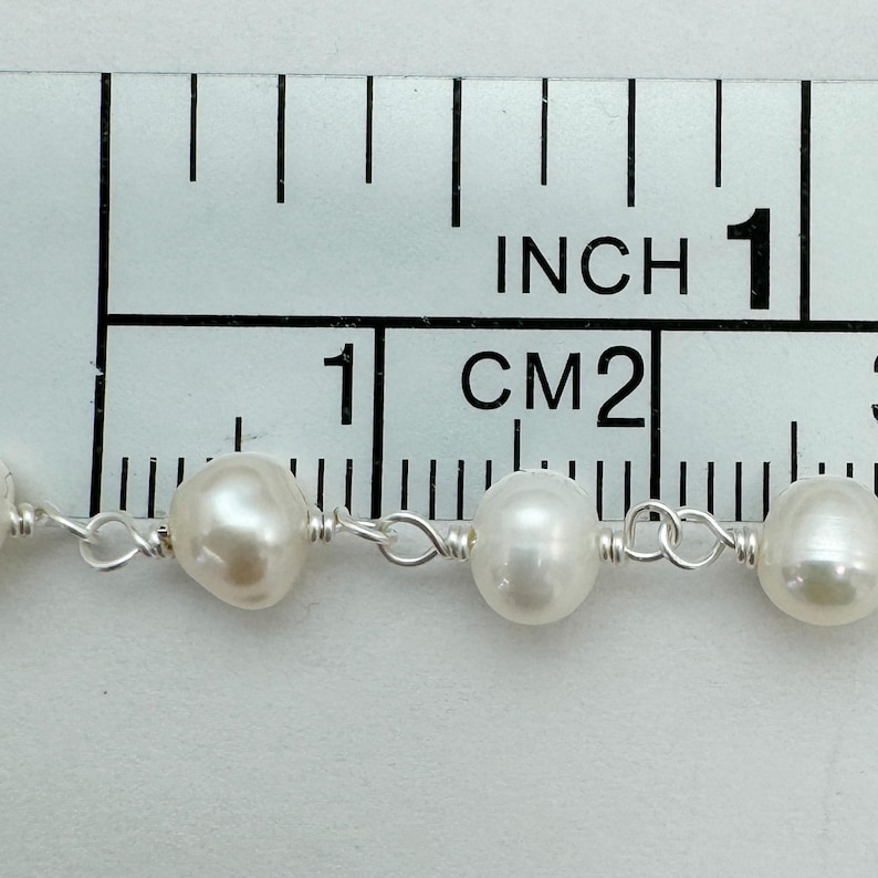 Puede incluir: Primer plano de un collar de perlas blancas de agua dulce, cada una de aproximadamente 1,3 cm de di&aacute;metro, ensartadas en un delicado alambre de plata. Las perlas se miden contra una regla marcada en pulgadas y cent&iacute;metros.