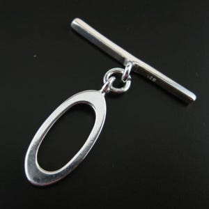 Puede incluir: Cierre de palanca de plata con un bucle redondo de forma ovalada. La palanca es una barra recta con un extremo redondeado.